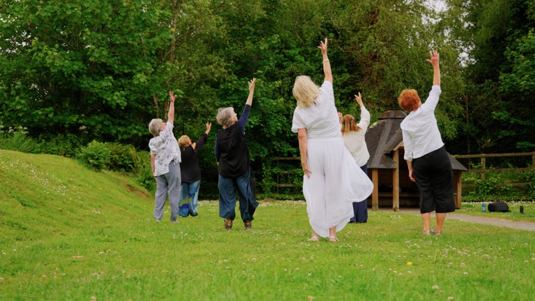 Members of Hayle WI creating for Wildworks' 'This is Me'. 6 people stand barefoot on grass with their back to the camera and their right hands raised